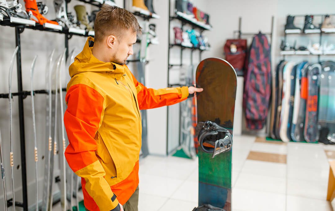 A close-up of a man in a yellow and orange jacket checking a snowboard inside a ski rental store.