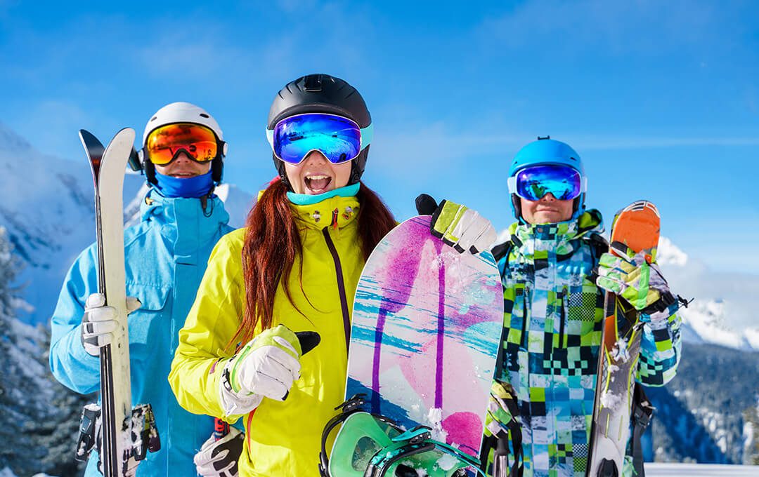 A close-up of three people holding a snowboard and ski blades, wearing skiing equipment, against a snowy mountain in the background