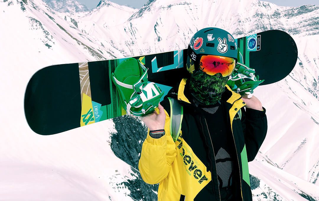 A close-up of a skier wearing yellow and green equipment carrying a snowboard against a snowy mountain background
