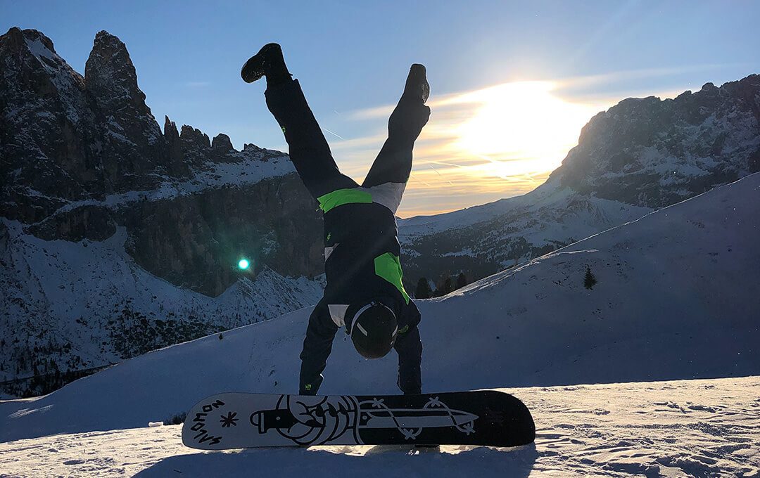 A man does a handstand in front of a ski board against a backdrop of mountains and a bright sky in the background.