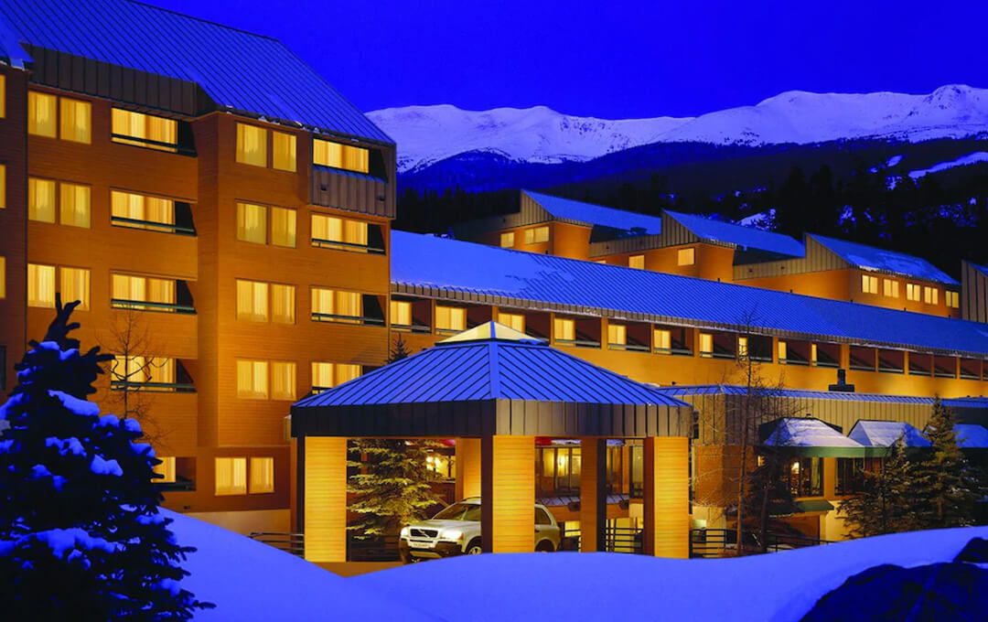 A front view of a hotel at the mountain base, surrounded by trees and snow, with a mountain range in the background.