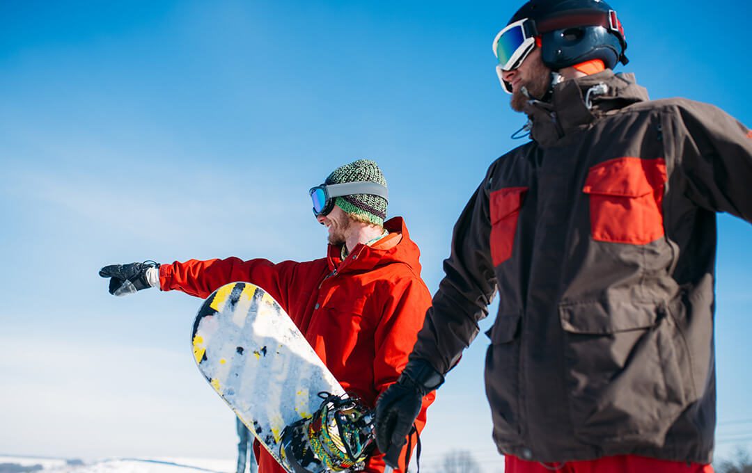 A close-up of two men wearing helmets, goggles, and outer jackets The man in an orange outer jacket was holding a snow board and pointing his finger at the slope of a mountain.