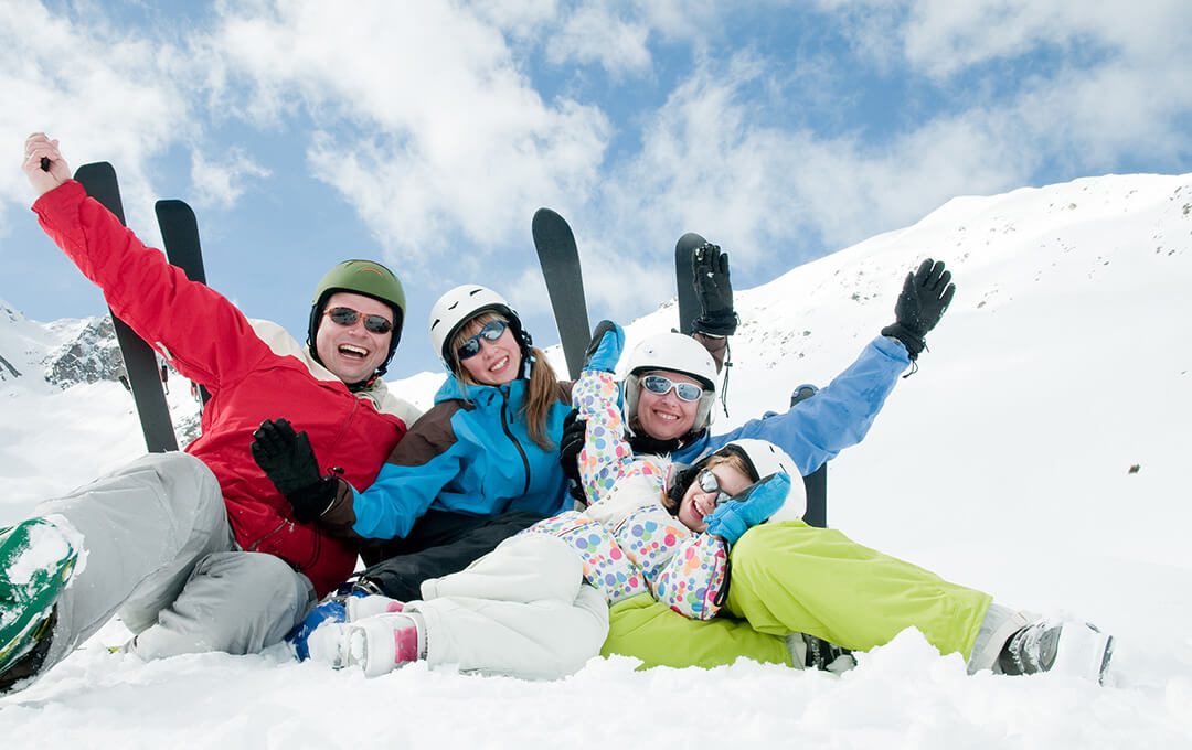 A close-up of a family wearing skiing gear sitting on snow against a snowy mountain in the background.