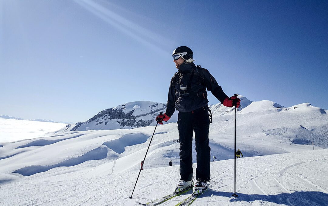 A close-up of a man wearing ski equipment standing on a ski track against a snowy mountain in the background.