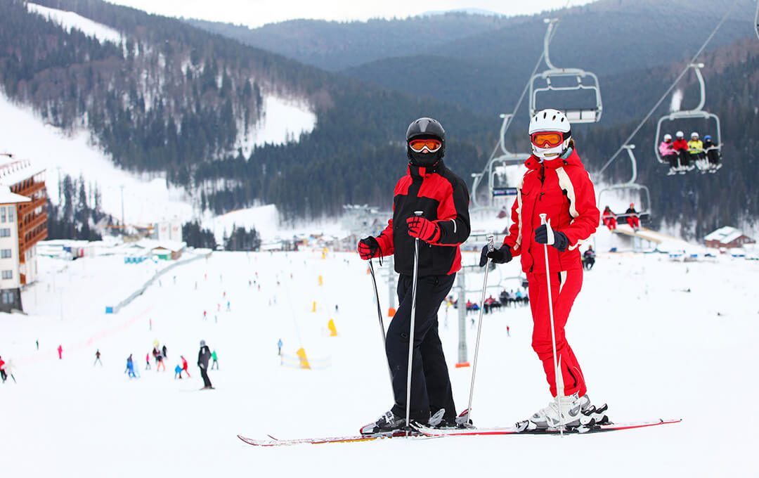 A wide shot of two people standing on a ski park against a snowy mountain with pine trees, cable cars with people, and people skiing in the background.