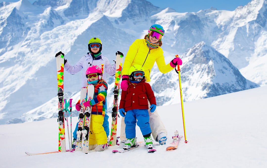 A full shot of a mother and three children wearing skiing gear standing on snow against a snowy mountain in the background.