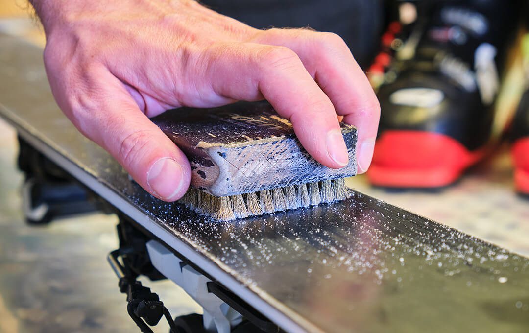 A close-up of a hand using a brush to apply wax to a ski blade.